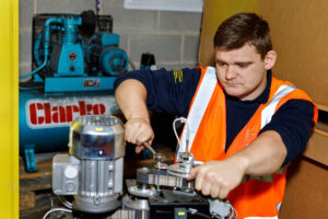 Worker in a high-visibility orange vest operating a belt-driven metal machine in a workshop.