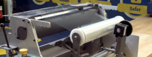 Close-up of a roller-based industrial machine with a white rolling belt and metal frame on a wooden worktable in a workshop setting.